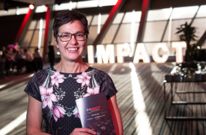 A photo of Kathryn standing in front of an sign that says IMPACT and holding a brochure in her hands